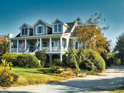 The front of the house in morning sun: a raised two-storey shingled house with three dormers, a covered porch across the front, two flags flying, and clipped shrubs along the drive. Nothing out of place.